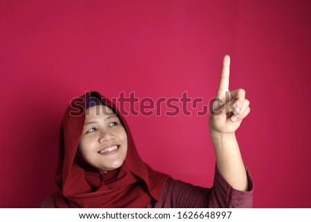 Portrait of Asian muslim woman smiling happily with finger pointing or touching virtual screen against red background, number one sign copy space template
