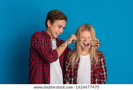 Sibling relationship concept. Elder brother teasing little sister, pulling her ears till she scream, standing together over blue studio background Royalty-Free Stock Photo #1626677470