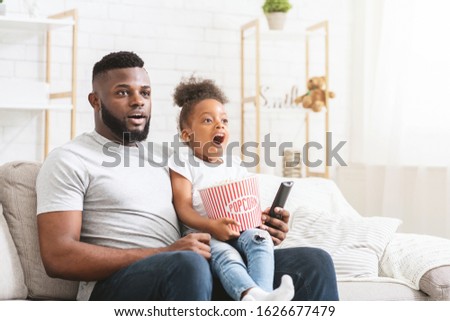 Emotional black father and preschool daughter watching action movies together at home, eating popcorn, free space