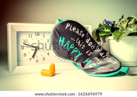 A modern clock with an alarm clock in white, a funny sleep mask, earplugs and a flower in a pot on the nightstand near the bed in the bedroom close-up. Old vintage retro grunge style photo.