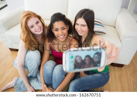 Happy young female friends photographing themselves with smartphone on floor at home
