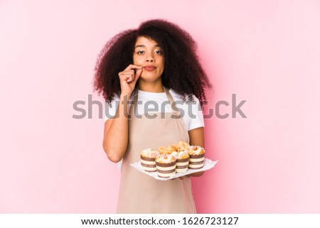 Young afro pastry maker woman holding a cupcakes isolatedYoung afro baker woman with fingers on lips keeping a secret.
