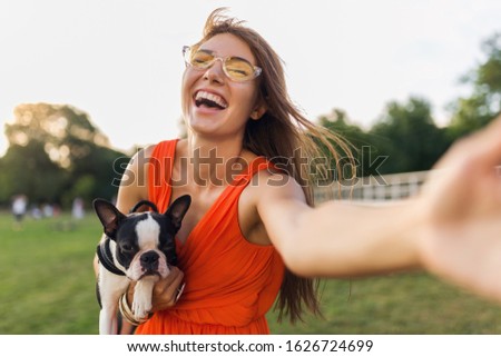 young happy smiling woman in orange dress having fun playing with dog in park, summer style, cheerful mood, making selfie photo on camera