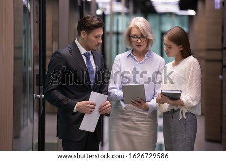 Concentrated middle aged blonde female team leader in eyeglasses holding computer tablet, showing product info, project results, mistakes in software to focused younger colleagues in office corridor.