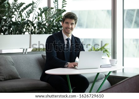 Confident young smiling successful businessman employee sitting on workplace with laptop, looking at camera. Happy manager salesman broker financial advisor in suit posing for photo in modern office.