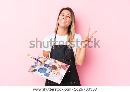 Young artist caucasian woman holding a palett isolated joyful and carefree showing a peace symbol with fingers.