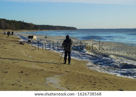 The shore of the Bay, yellow sand, gray stones, white ice on blue water, the water's edge is powdered with snow, blue sky.People walk along the shore, the photographer takes a picture of the landscape