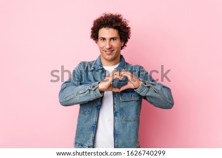 Curly mature man wearing a denim jacket against pink background smiling and showing a heart shape with hands.