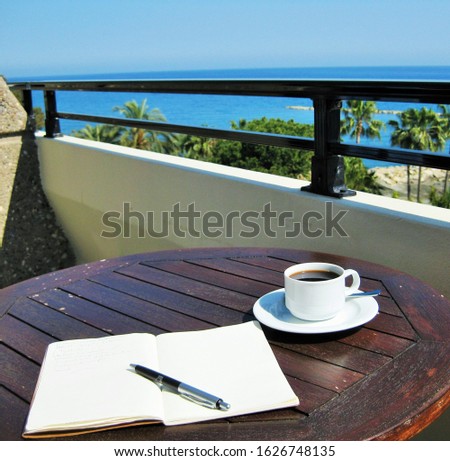 writer's place: open notebook with pen and cupof coffee on balcony table with blue sea in the background