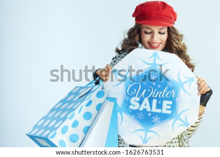 smiling modern woman with long brunette hair in sweater, scarf and red hat with shopping bags looking at winter sale banner on winter light blue background.