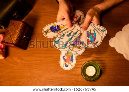 Hands of a mature woman holding divination cards. Pictures are hand-drawn based on old medieval fortune-telling cards. Fortunetelling cards, candles, magic attributes on the wooden background.