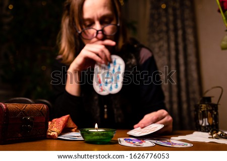 Faddish middle-aged fortuneteller with round glasses holds a card  in her hand.  Selective focus on the candle in front,  image of a woman on the background in blur.