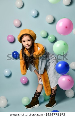 portrait of a little Asian woman in a denim dress and a stylish scarf with a beret., with a wide toothless smile against the background of many multi-colored balloons. Birthday Party, Fashion