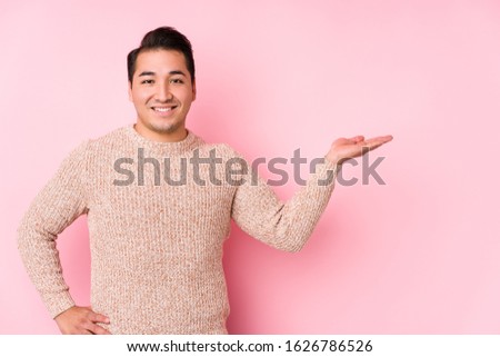 Young curvy man posing in a pink background isolated showing a copy space on a palm and holding another hand on waist.