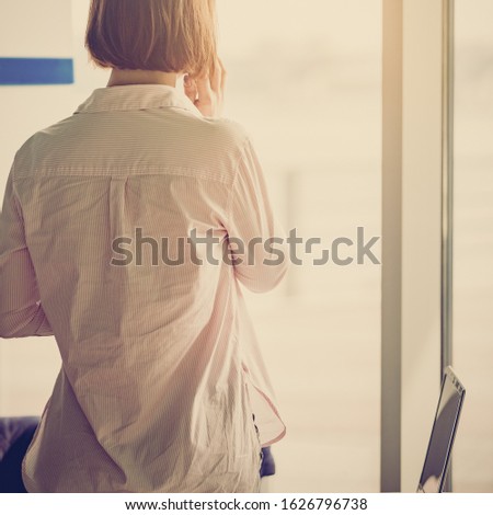 Successful businesswoman talking on phone with client and looking on window, back view photo