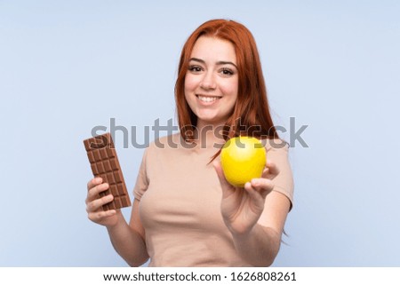 Redhead teenager girl over isolated blue background taking a chocolate tablet in one hand and an apple in the other