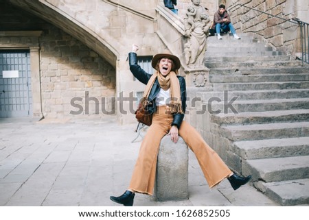 Pretty hipster female traveler with backpack wearing trendy clothes having fun while sitting like cowboy with raised hand on stone next to stairs of old building and screaming