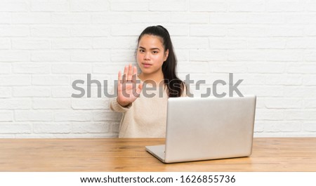 Young teenager Asian girl with a laptop in a table making stop gesture with her hand