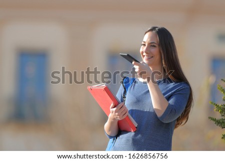 Happy student using voice recognition on cell phone standing outdoors in a university campus