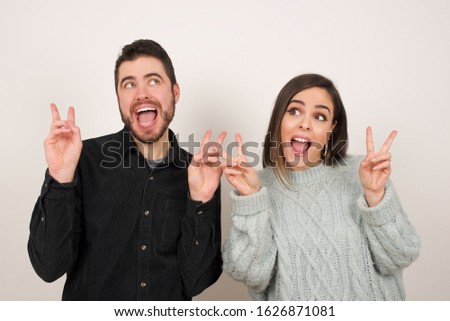 Isolated shot of cheerful couple makes peace or victory sign with both hands, dressed in casual clothes, feels cool has toothy smile, isolated over gray background. People and body language.