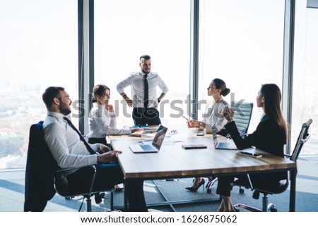 Group of elegant business people sitting at table listening to speaker brainstorming and using laptops during meeting in modern glass office