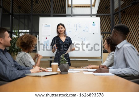 Smiling millennial Asian female coach or speaker stand hold company team meeting or making presentation for employees, confident ethnic woman speak presenting business project for colleagues Royalty-Free Stock Photo #1626893578