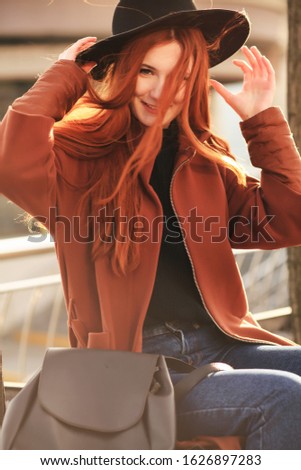 Young red haired girl in winter coat standing outside and watching the sunset in the city, the sun shines brightly, fashionable clothes on a girl, hipster style, backpack, sun, street photo, mood. 