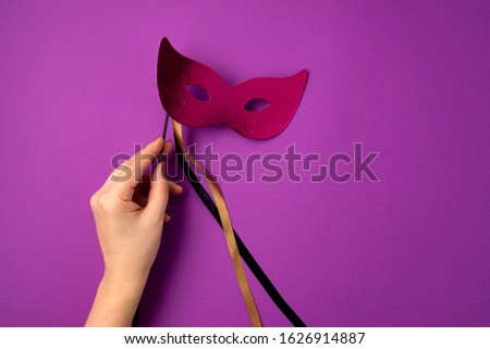 Woman hand holding  festive, colorful mardi gras or carnivale mask over purple background. Party invitation, greeting card, venetian carnivale celebration concept. Flat lay, top view