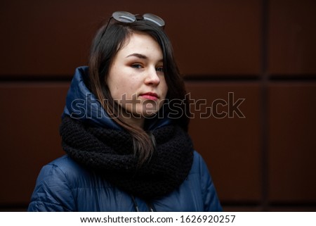 Young pretty white caucasian girl with problem skin in a blue jacket and glasses opposite modern brown background on the street in the winter.