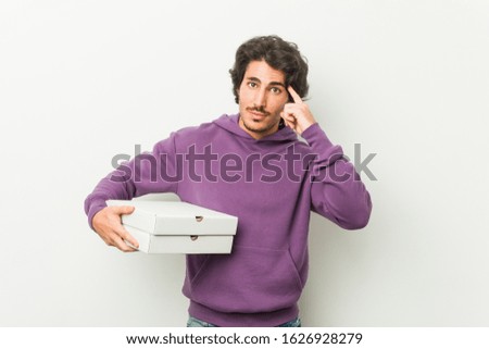 Young man holding pizzas package pointing his temple with finger, thinking, focused on a task.