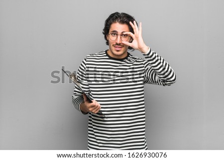 Young employee man holding an inventory showing okay sign over eyes