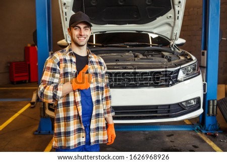 smiling male technician looking at the camera showing thumb up in the garage with car on the background