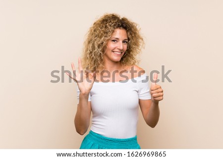 Young blonde woman with curly hair over isolated background showing ok sign and thumb up gesture