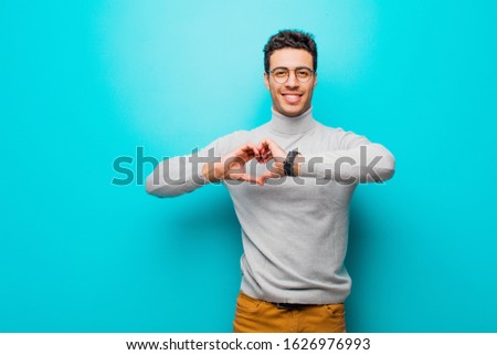 young arabian man smiling and feeling happy, cute, romantic and in love, making heart shape with both hands against flat wall