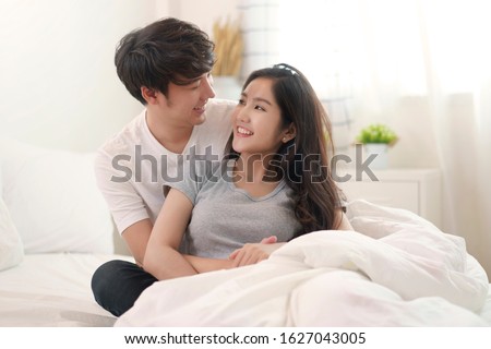Asian young couple man and woman staying together in bedroom. Husband hugging wife from behind, looking eyes at each other feeling lover happy with smile face on morning. Relationship of love concept.