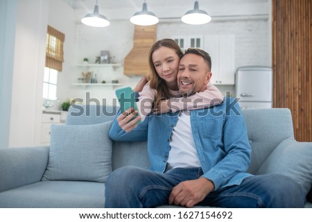 Selfie with dad. Happy daughter hugging the neck of the father, who is sitting on the couch, photographing himself with his father on a smartphone.