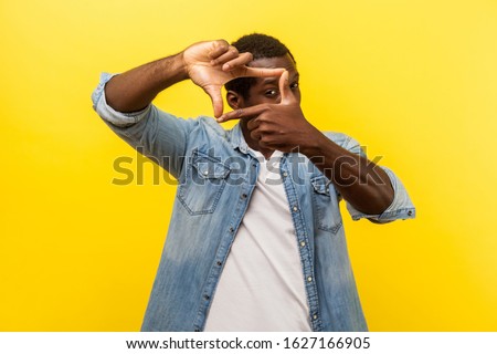 Portrait of focused curious man in denim casual shirt with rolled up sleeves looking through photo frame made of hands, viewing distant with interest. indoor studio shot isolated on yellow background