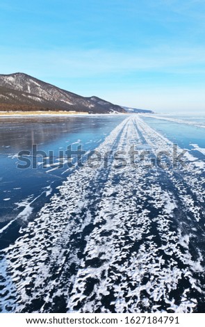 Baikal Lake on a cold day in January. The ice road from Listvyanka to the village of Goloustnoye with snow tracks from Khivus on the surface of blue ice. Beautiful winter landscape. Ice travel
