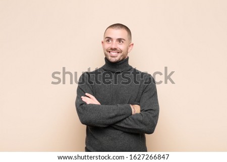 young handsome man feeling happy, proud and hopeful, wondering or thinking, looking up to copy space with crossed arms against flat wall