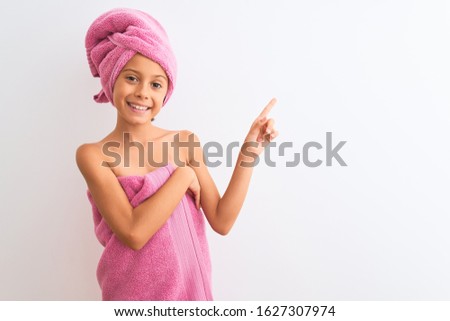 Beautiful child girl wearing shower towel after bath standing over isolated white background with a big smile on face, pointing with hand and finger to the side looking at the camera.
