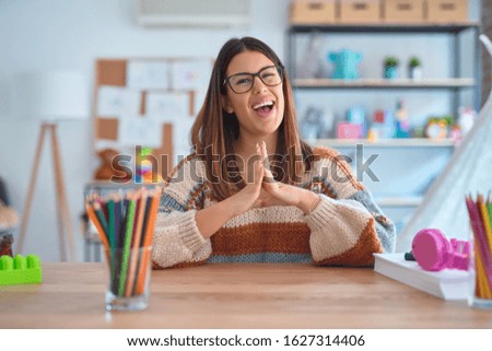 Young beautiful teacher woman wearing sweater and glasses sitting on desk at kindergarten Hands together and fingers crossed smiling relaxed and cheerful. Success and optimistic