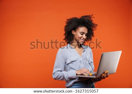Attractive young african woman wearing casual clothes standing isolated over red background, using laptop computer