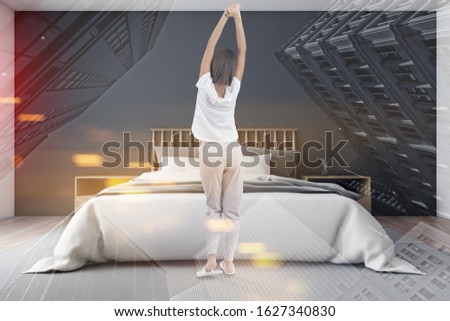 Rear view of woman in pajamas stretching in luxury bedroom with gray walls, and wooden king size bed. Concept of good morning. Toned image double exposure