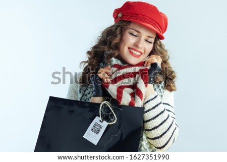 happy young 40 years old woman with long brunette hair in sweater, scarf and red hat with shopping bags enjoying purchased sweater isolated on winter light blue background.