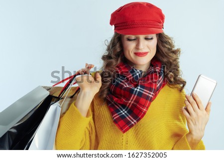 relaxed elegant female with long brunette hair in sweater, scarf and red hat with shopping bags and smartphone on winter light blue background.