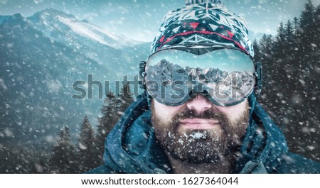 Close-Up Portrait of Caucasian holiday man with beard in winter snow landscape. Beautiful mountains view reflected in ski glasses. Sunny frozen day.