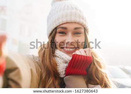 Close up of a cheerful pretty young girl wearing winter jacket and a hat standing on a city street, taking a selfie