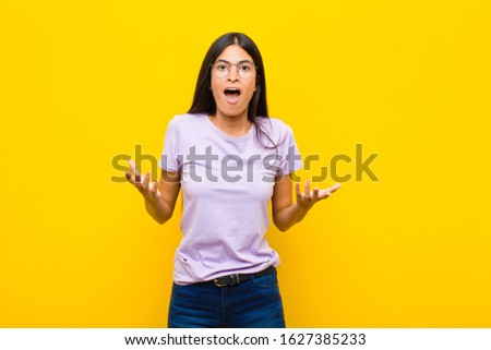 young pretty latin woman feeling extremely shocked and surprised, anxious and panicking, with a stressed and horrified look against flat wall