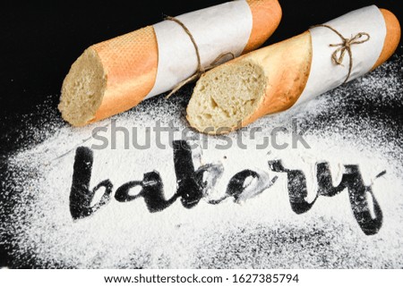 Inscription bakery on white wheat flour scattered Sliced french baguette on dark background, hand written in flour the word bakery