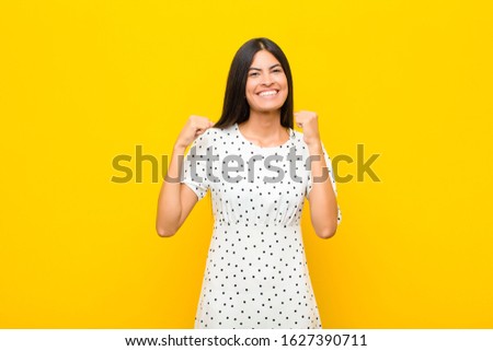 young pretty latin woman feeling happy, positive and successful, celebrating victory, achievements or good luck against flat wall